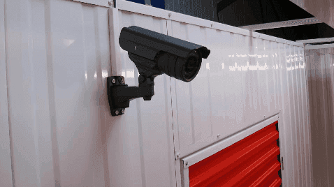 Black surveillance camera mounted on white metal wall above a red storage unit door at HOMEBOX Braga